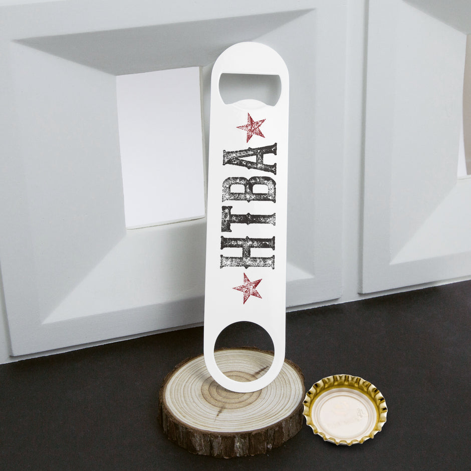A white bottle opener with 'HIBA' in distressed black letters and two red stars, displayed on a wood slice coaster next to a gold bottle cap on a dark surface.