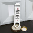 A white bottle opener with 'HIBA' in distressed black letters and two red stars, displayed on a wood slice coaster next to a gold bottle cap on a dark surface.
