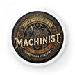 A circular metal cap or bottle stopper featuring a vintage-style machinist design. The central label has a black background with ornate gold and beige typography reading 'MACHINIST' and 'FINE PRECISION & QUALITY CRAFTSMANSHIP ENGINEERING & METALWORK,' surrounded by illustrated gears, tools, and stars.
