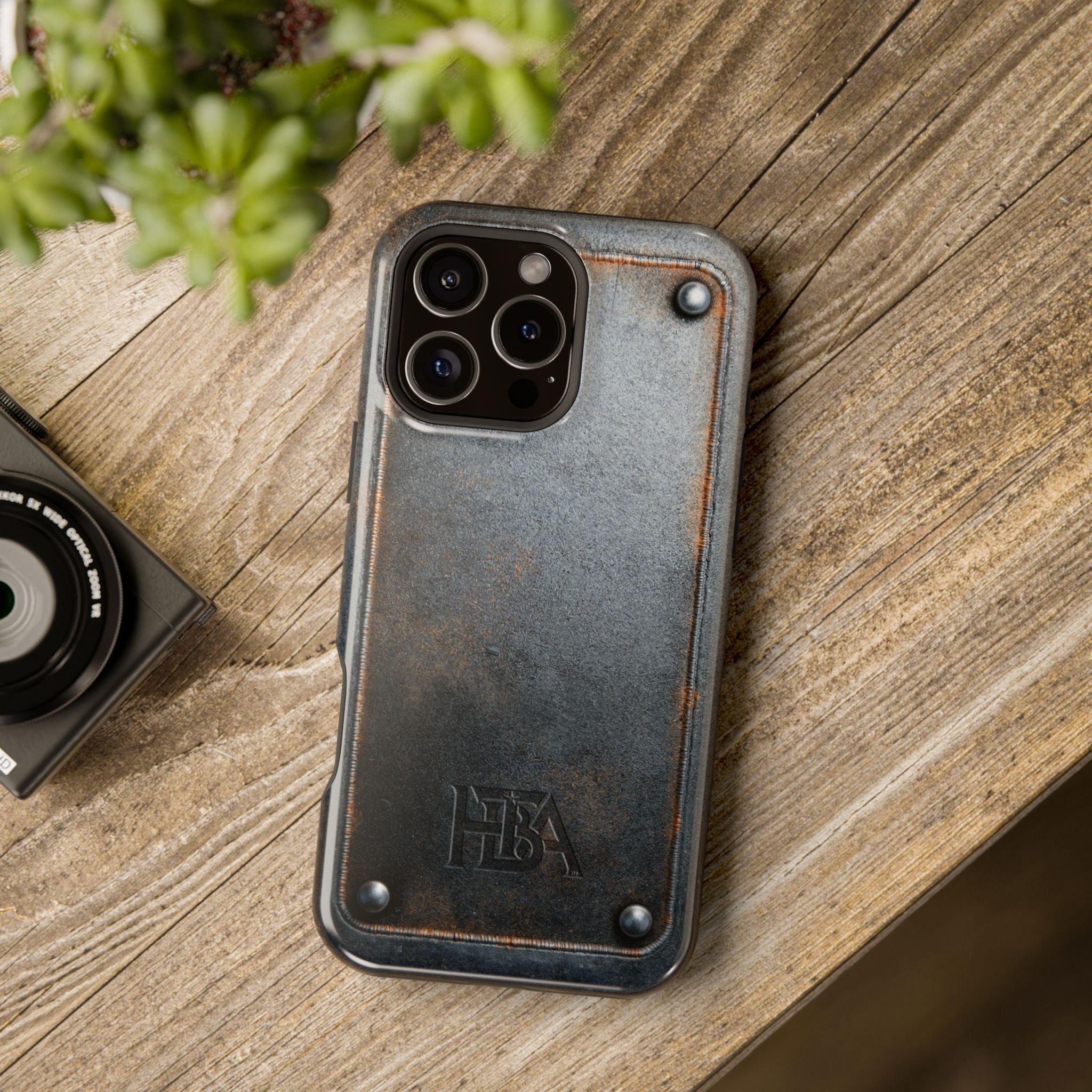 Phone case with 'HBA' logo on a wooden surface next to a camera and plant