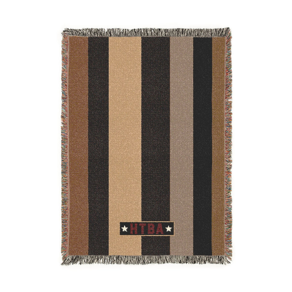 A rectangular woven throw blanket with horizontal stripes in brown, black, tan, and beige, finished with fringed edges and a vertical black brand tab reading 'KILTI' with two white stars.