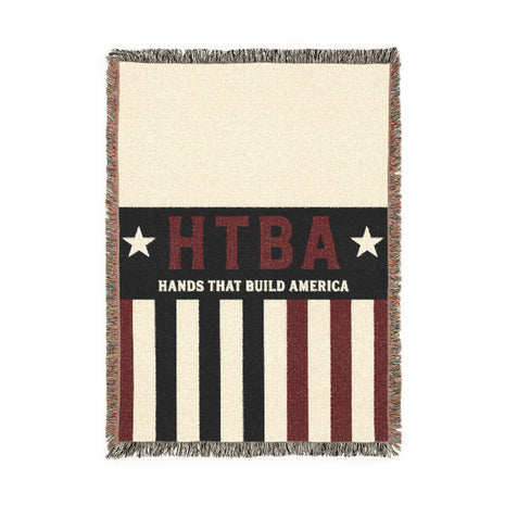 A rectangular woven throw blanket with fringe edges, featuring a patriotic American flag-inspired design. The left side is cream, the center has a black vertical band with red 'HTBA' lettering, white stars, and white text reading 'HANDS THAT BUILD AMERICA', and the right side has alternating cream and dark red stripes.