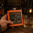 A hand holds an orange square flask with a detailed black and gold 'Ironworker' graphic, set on a rustic wooden table next to a glass of whiskey, with a warm, industrial-themed background.