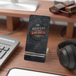Phone stand with 'Roofers That Build America' logo on a desk with computer and headphones.