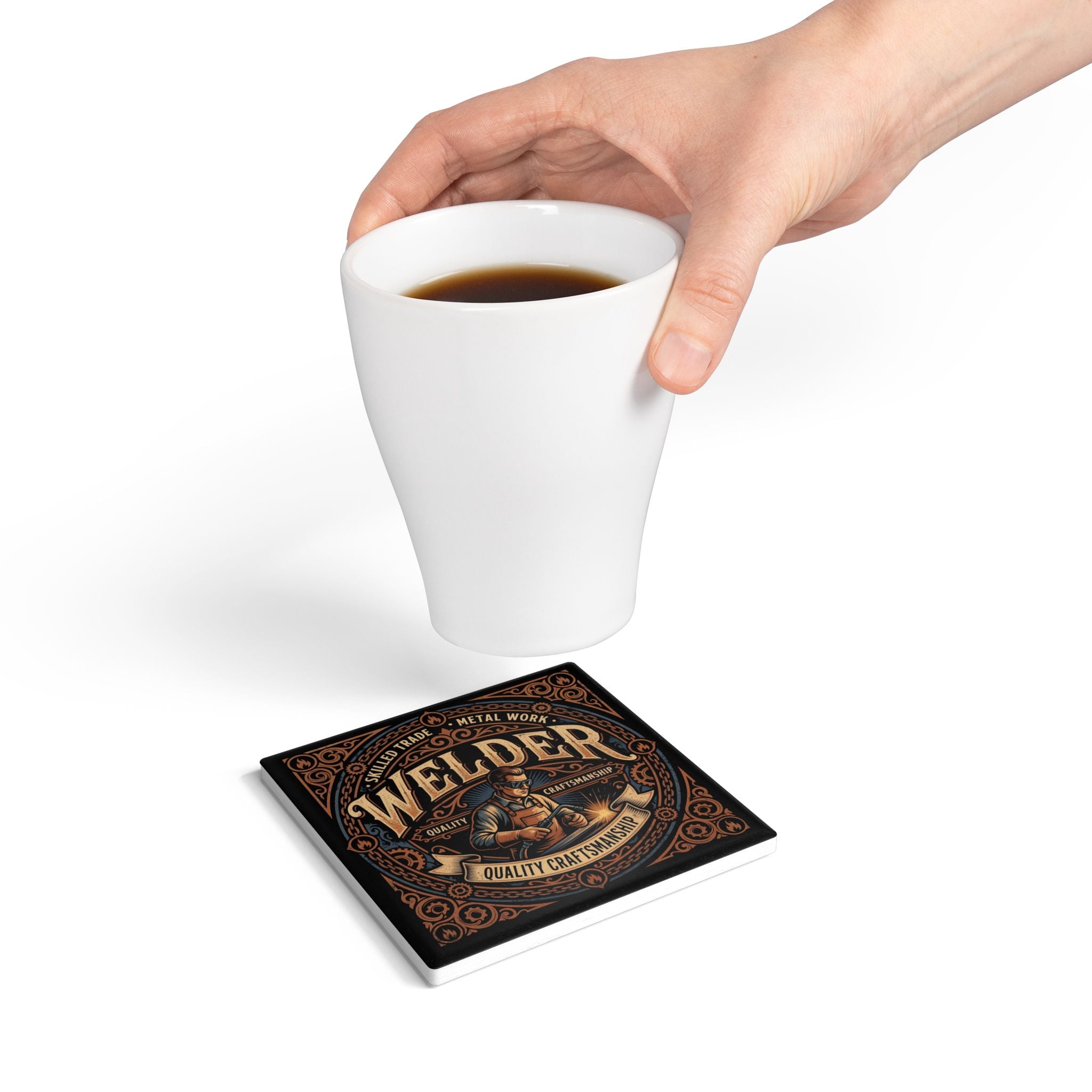 A square ceramic coaster with a vintage-style 'Welder' graphic is shown on a white surface, with a hand holding a white coffee mug above it.