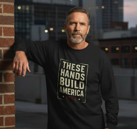 Man wearing a black long-sleeve shirt / jersey with 'These Hands Build America' and red HTBA. Leaning against a brick wall Inner city. 
