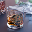 A clear rocks glass with a printed welding-themed badge that reads “WELDER” and smaller text “WELDING-FABRICATION-MAINTENANCE,” filled with amber liquid, set on a dark tabletop with blurred background objects.